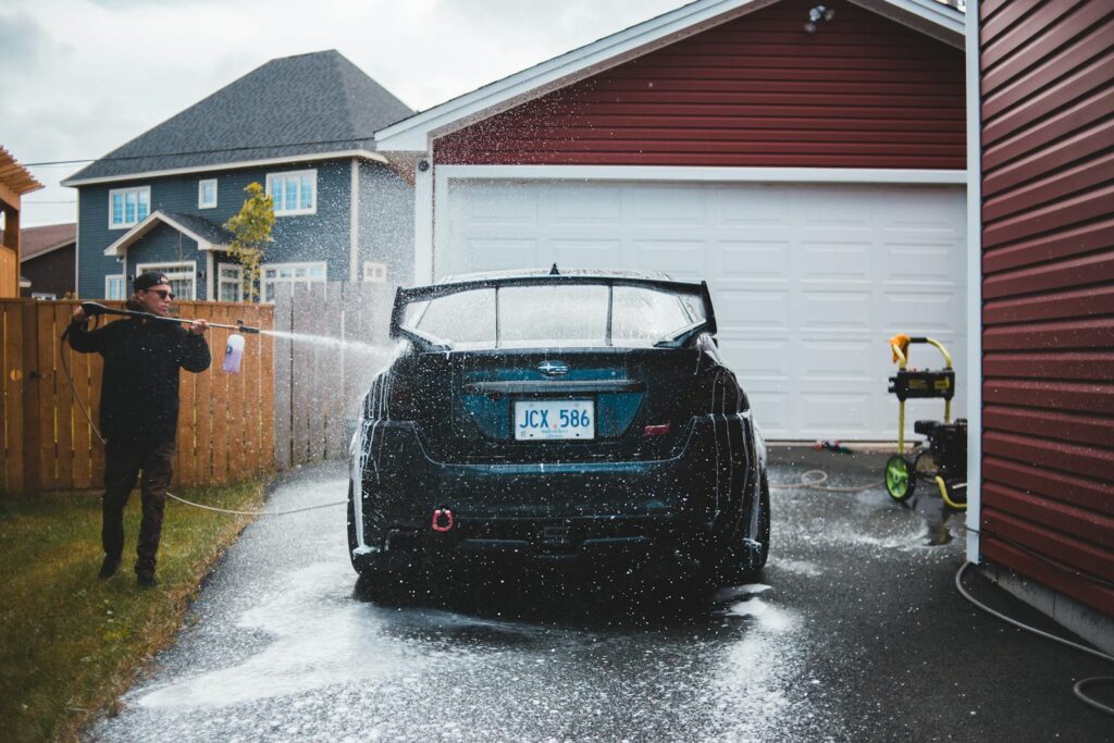 A man uses a hose to wash a sports car in a residential driveway, creating a splash effect.
