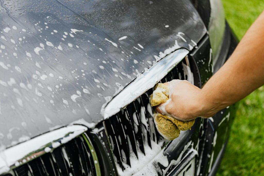Detailed view of a hand washing a car grille with a soapy sponge outdoors.