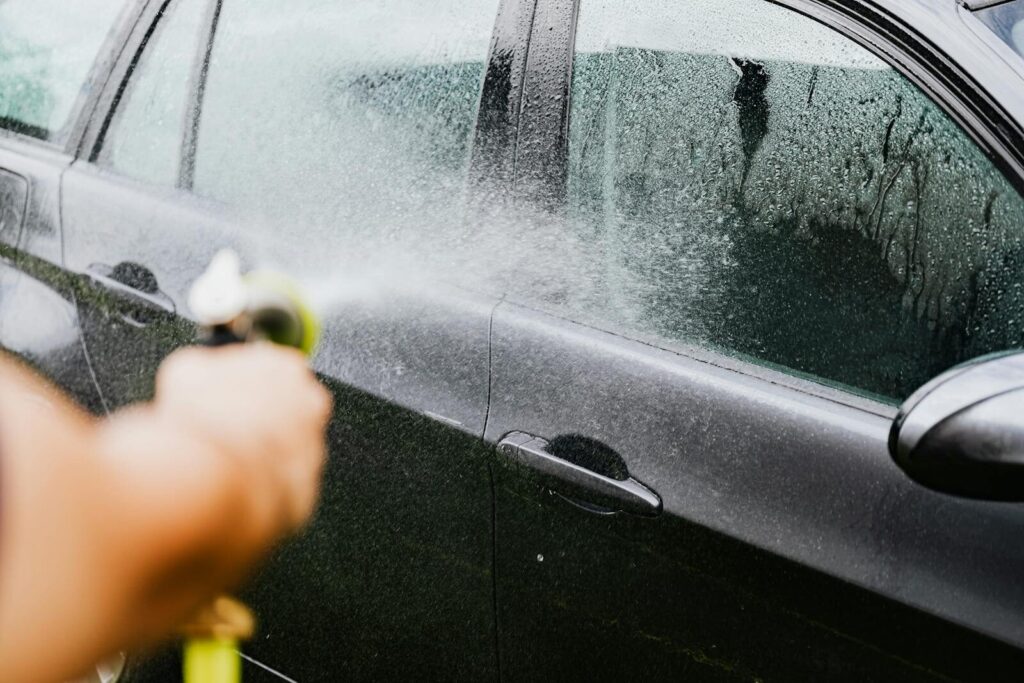 A car being washed with a hose outdoors. Water sprays create a cleaning effect on the vehicle.