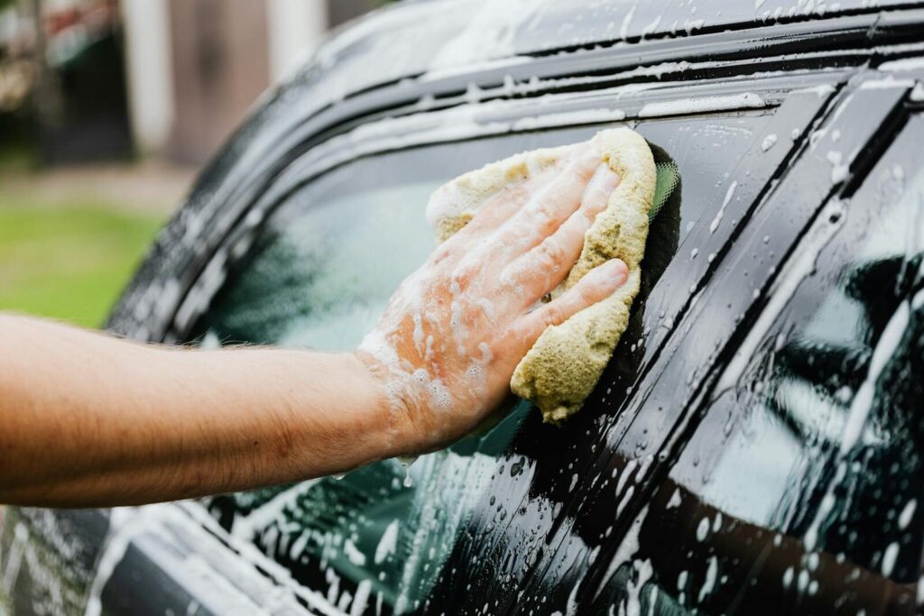 A hand scrubs a car window with a sponge, covered in soap suds, during a car wash.
