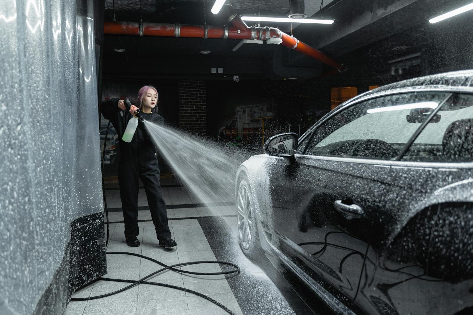 Photo by Tima Miroshnichenko Woman using a pressure washer to clean a black car in an indoor car wash setting.