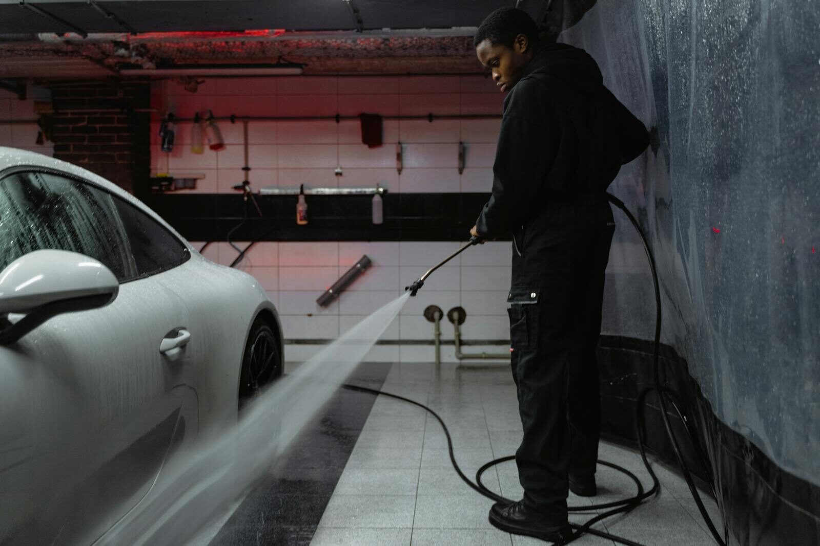 Photo by Tima Miroshnichenko African American man cleaning a white car with a pressure hose in an indoor garage setting.