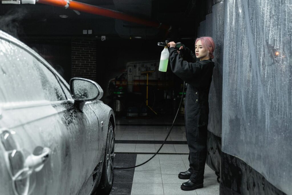 A young woman uses a power spray to clean a car with foam in an indoor garage.