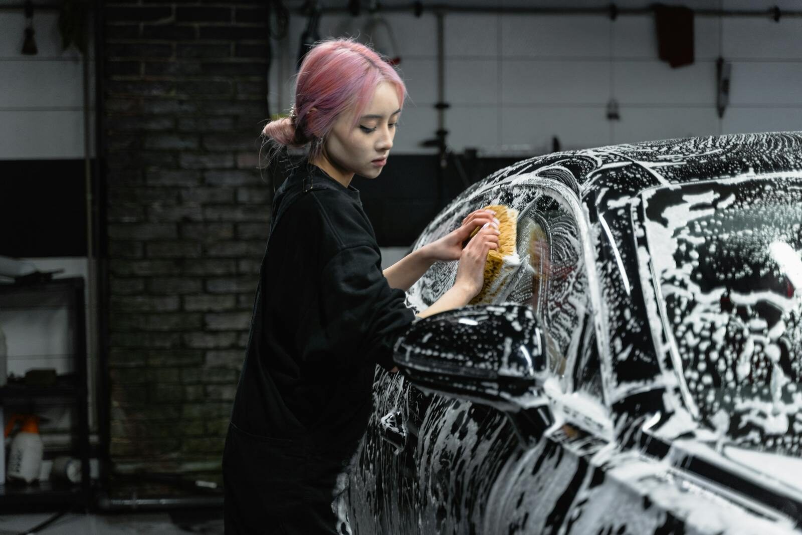 Woman with pink hair washing a car in a garage using soap and a sponge.
