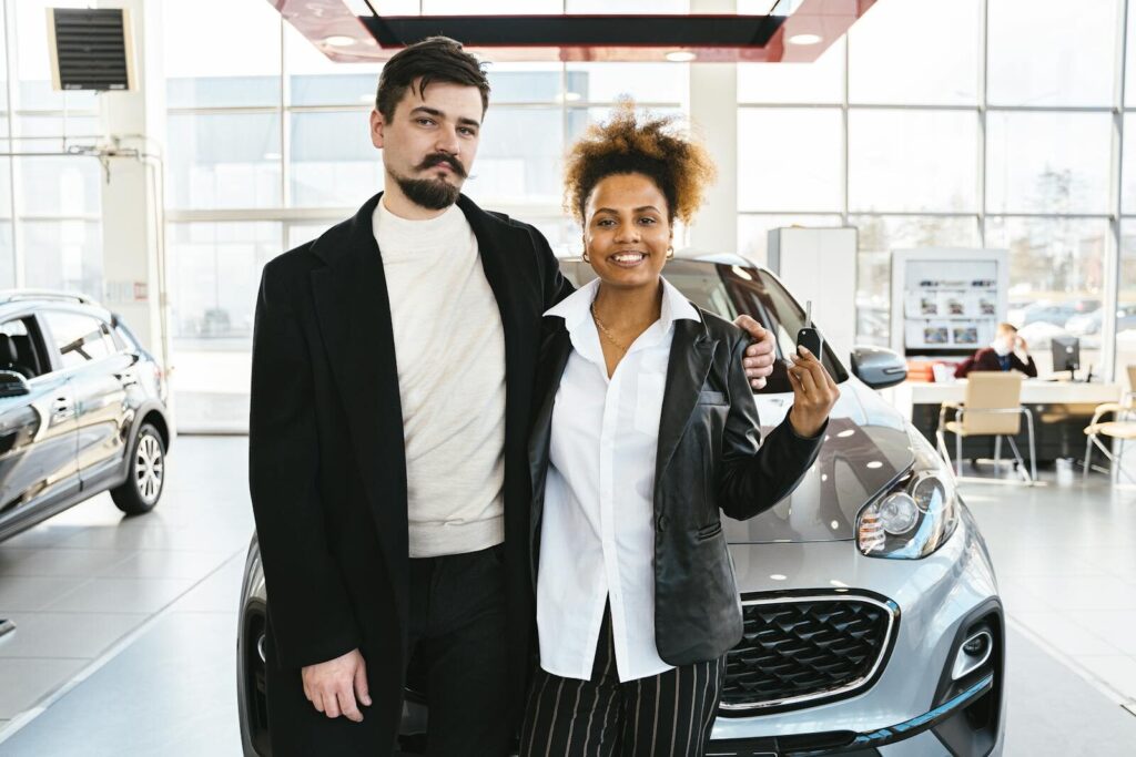 Interracial couple celebrates buying a new car at a dealership, holding keys and smiling.